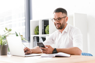 Photo of smiling adult man 30s in white shirt using laptop and mobile phone while sitting at table and working in office - Powered by Adobe