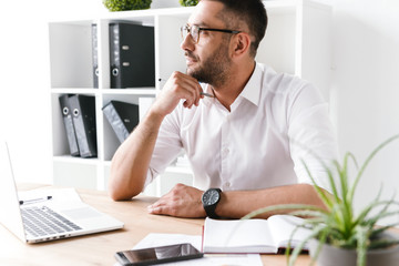 Image of concentrated businessman 30s in white shirt looking aside and thinking while writing down...