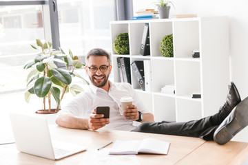 Image of cheerful handsome man in formal wear sitting in office with legs at table and using black smartphone