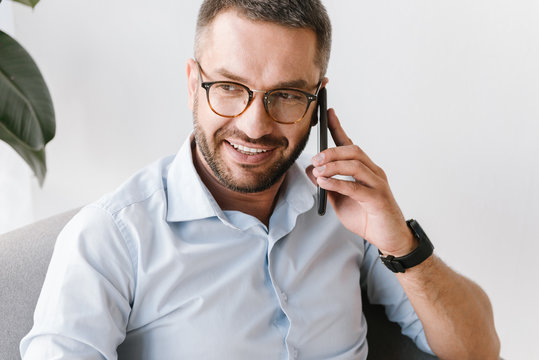 Image closeup of handsome man in white shirt smiling and talking on black smartphone about business, while working in office