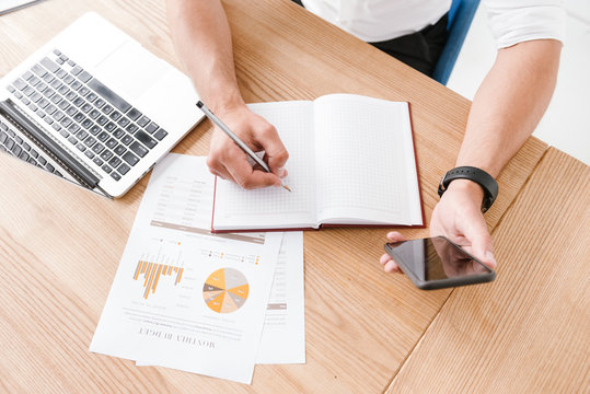 Cropped Image From Above Of Caucasian Man Sitting At Table And Writing Down Data Or Report At Notebook From His Mobile Phone, While Working In Bright Office Room