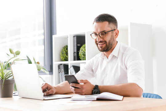 Photo of smiling adult man 30s in white shirt using laptop and mobile phone while sitting at table and working in office