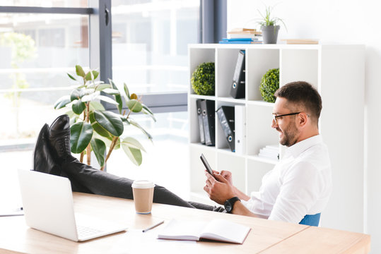 Image Of Happy Handsome Man In Formal Wear Sitting In Office With Legs At Table And Using Black Smartphone