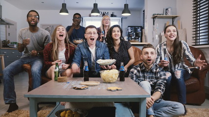 Diverse group of friends watching sports on TV Football supporters celebrate success with popcorn...
