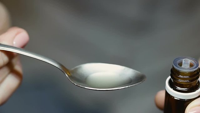 Lady pours ten drops of turbid white liquid out of bottle on teaspoon and drinks