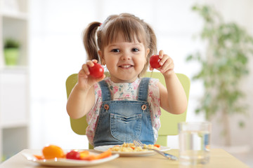 Healthy kids nutrition concept. Cute toddler girl sitting at table with plate of salad, vegetables, pasta in room. Child eating healthy food.