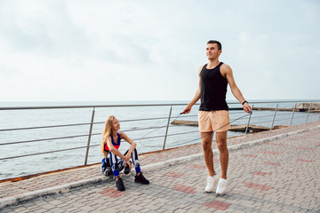 Young sportive couple working out together on the quay, near the sea. Handsome man jumping with skipping rope, while his girlfriend watching at him and smiling. - obrazy, fototapety, plakaty