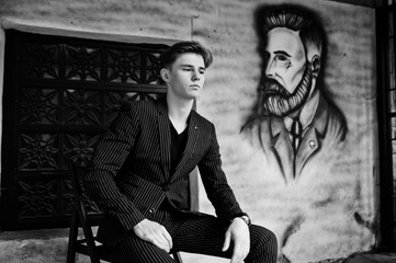 Young stylish macho boy in black jacket posed outdoor of street. Amazing model man sitting against grafiti wall of barbershop.