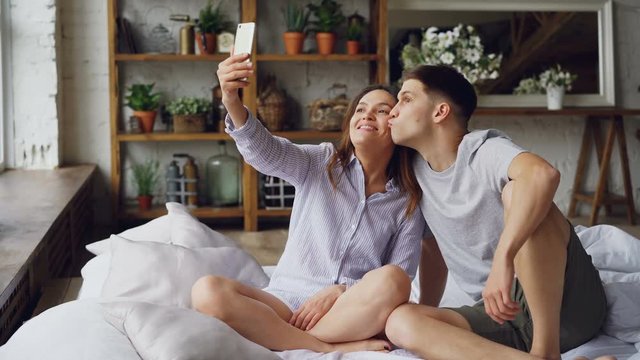 Cheerful loving couple is taking selfie with smartphone looking at camera, posing and making funny faces while sitting together on bed at home. Technology and relationship concept.