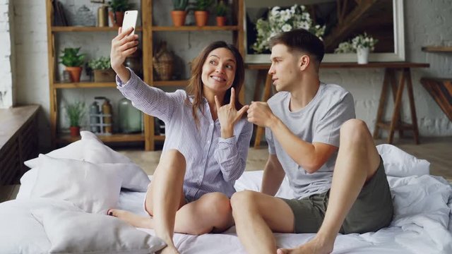 Modern married couple is taking selfie in bedroom gesturing posing and kissing while sitting on bed together. Modern technology and people concept.