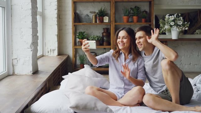 Young woman is holding smartphone, touching screen and making video call together with her husband while sitting on bed at home. Communication and people concept.