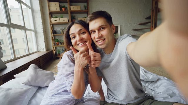 Point of view shot of loving couple taking selfie together posing, kissing and having fun while sitting on bed at home. Nice modern interior and large windows are visible.