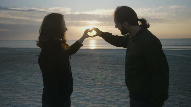 Beautiful couple making heart shape with their hands on the beach at sunrise and then holding their hands and kissing
