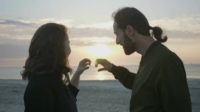 Modern couple enjoying sunset at the beach and making a heart shape with their hands while looking at the sun and then kissing romantically
