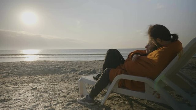 Beautiful young couple covered with a blanket hugging and lying on a lounge chair on the beach enjoying the sunset