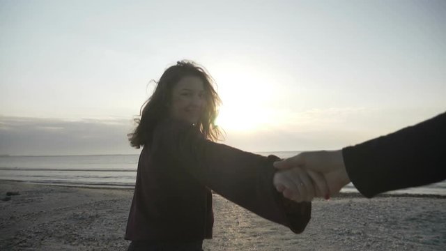 Young couple holding hands girl leading boyfriend walking towards sea sunrise on empty beach POV travel concept in slow motion