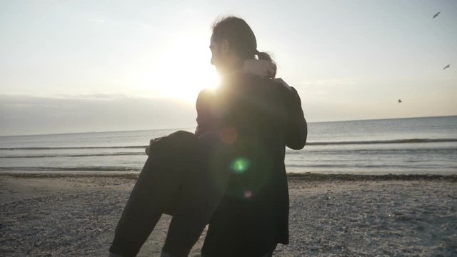 Young happy couple in love swirling on the beach man holding woman in his arms while sun goes down at sunset in slow motion
