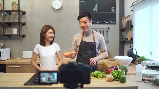 Young asian couple man and woman in kitchen recording video on camera. Smiling asian couple working on food blogger concept with fruits and vegetables in kitchen.