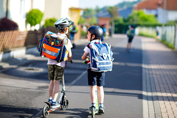 Two school kid boys in safety helmet riding with scooter in the city with backpack on sunny day....