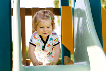 Happy blond little toddler girl having fun and sliding on outdoor playground. Positive funny baby...