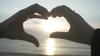 Couple making heart shape with their hands with sunset inside on the beach in slow motion