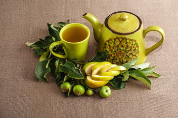 tea pot, a cup of tea, branch with unripe small green apples and one big curly cutting apple on the sack background