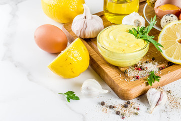 Homemade mayonnaise sauce with ingredients - lemon, eggs, olive oil, spices and herbs, white marble kitchen table copy space
