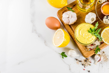 Homemade mayonnaise sauce with ingredients - lemon, eggs, olive oil, spices and herbs, white marble kitchen table copy space