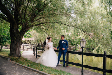 The husband leads his wife by the hand in the park with a pond, and shows beautiful places. The bride in a wedding dress follows her man in a stylish suit.