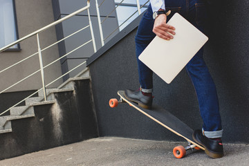 cropped shot of young man holding laptop and riding skateboard