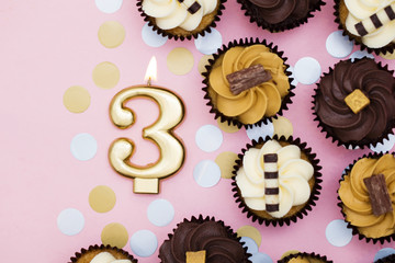 Number 3 gold candle with cupcakes against a pastel pink background