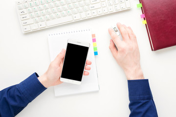 Desktop, male hands, man working at the computer. Guy holding a white smart phone, white notebook. Background with copy space, for advertisement, top view