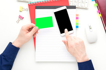 white desktop, male hands, man holding a business card and uses smart phone, red notebook, white background with copy space, for advertisement, top view
