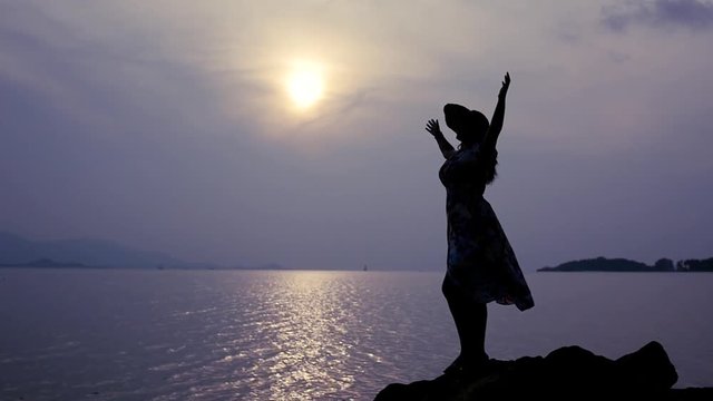 A lonely girl stands on the rocks on the beach, admires the evening and the sunset, lifts her hands up, feels freedom. slow motion, 1920x1080, full hd