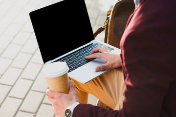 cropped shot of man holding paper cup and using laptop with blank screen on street