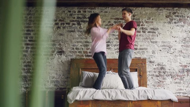 Young husband and wife in casual clothes are jumping and dancing on bed, laughing and having fun in nice loft style apartment. People and entertainment concept.