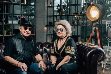 Fototapeta na wymiar Happy couple of retirees in biker clothes.pair of seniors in stylish black clothes sitting on a leather couch.Senior man in black leather jacket and goggles.Unusual retired couple.