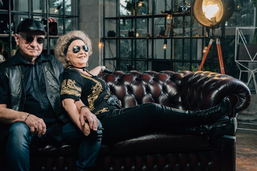 Happy couple of retirees in biker clothes.pair of seniors in stylish black clothes sitting on a leather couch.Senior motorcyclist with a black helmet and goggles looking at the camera.Happy pensioners