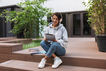 Beautiful smiling lady sitting in headphones and tablet in hands while spending time in courtyard of university