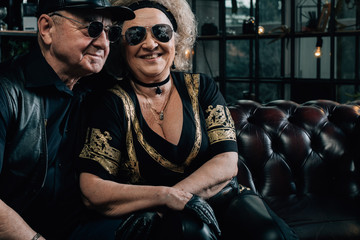 Fototapeta na wymiar Happy couple of retirees in biker clothes.pair of seniors in stylish black clothes sitting on a leather couch.Senior man in black leather jacket and goggles