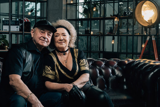 Happy couple of retirees in biker clothes.pair of seniors in stylish black clothes sitting on a leather couch.Senior man in black leather jacket and goggles