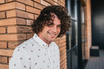 Horizontal closeup shot of smiling good-looking stylish male model with curly hair and freckles, wears trendy shirt, posing on outdoor building. People, lifestyle and business concept.