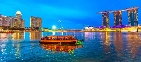 Fotobehang Panorama van Singapore gebouwen, wolkenkrabbers en reuzenrad weerspiegeld in de zee. Toeristische boot vaart & 39 s avonds in de baai. De horizon van Singapore bij blauw uur. Nachtscène waterkant jachthaven baai. © bennymarty