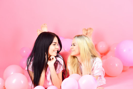 Girls lay on belly near balloons, pink background. Friendship concept. Sisters or best friends in pajamas at girlish pajamas party. Blonde and brunette on smiling faces have fun at domestic party.