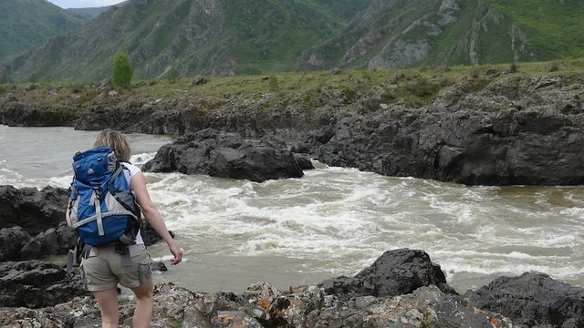 A girl with a backpack on her back goes to a mountain river, stands on a rock and looks at the stream.