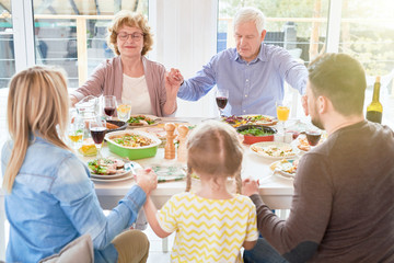 Portrait of big happy family joining hands in prayer at dinner holding hands during festive celebration in sunlight