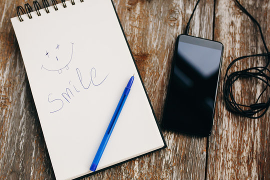 White notebook with a blue pen is lying on the old vintage background. Smile emotions is drawn and written on sketch book. Black smartphone with earphones is lying nearby. Modern technologies.