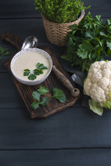 Cream of cauliflower soup with cream and greens, in a white cup on a wooden board and black background. Vintage photo. Fresh greens and cauliflower. Copy space