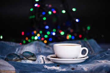 Cup with coffee and beans on plate on denim, blurred colorful lights on background. Mug full of black coffee and sunglasses on jeans, defocused. Festive coffee break concept