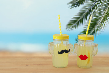 Image of fresh lemonade drink in cute cactus shape glasses wearing mustache and lips, over wooden table. Tropical summer romantic vacation concept.